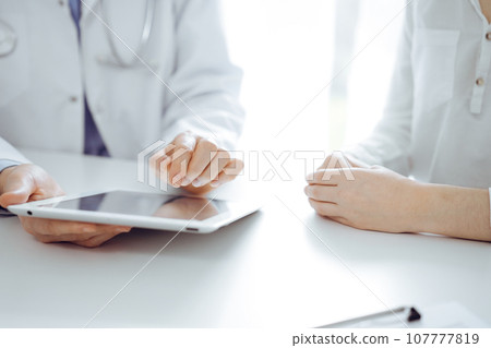 Doctor and patient sitting near each other at the desk in clinic. The focus is on female physician's hands pointing into tablet computer touchpad, close up. Medicine concept Doctor and patient sitting near each other at the desk in clinic. The focus is on female physician's hands pointing into tablet computer touchpad, close up. Medicine concept 107777819