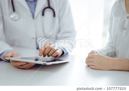 Doctor and patient sitting near each other at the desk in clinic. The focus is on female physician's hands pointing into tablet computer touchpad, close up. Medicine concept Doctor and patient sitting near each other at the desk in clinic. The focus is on female physician's hands pointing into tablet computer touchpad, close up. Medicine concept 107777820