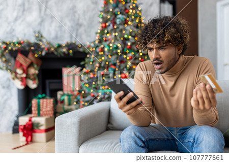 Cheated and upset Christmas man in living room near decorated Christmas tree, rejected and wrong money transfer, hispanic man holding bank credit card and phone. 107777861