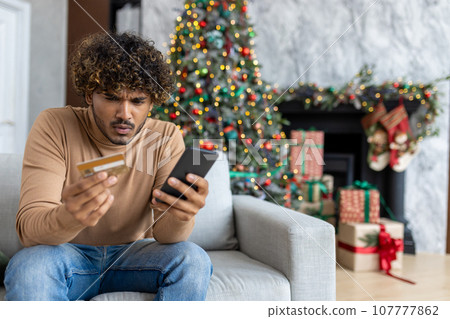 Cheated and upset Christmas man in living room near decorated Christmas tree, rejected and wrong money transfer, hispanic man holding bank credit card and phone. Cheated and upset Christmas man in living room near decorated Christmas tree, rejected and wrong money transfer, hispanic man holding bank credit card and phone. 107777862