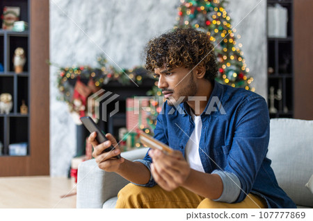 Cheated and upset Christmas man in living room near decorated Christmas tree, rejected and wrong money transfer, hispanic man holding bank credit card and phone. Cheated and upset Christmas man in living room near decorated Christmas tree, rejected and wrong money transfer, hispanic man holding bank credit card and phone. 107777869