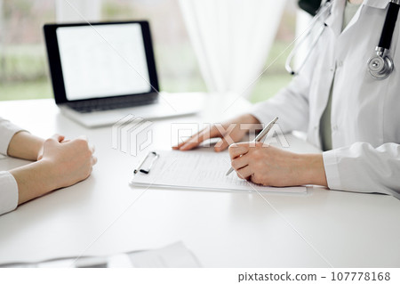 Doctor and patient sitting near each other at the white desk in clinic. Female physician is listening filling up a records form. Medicine concept Doctor and patient sitting near each other at the white desk in clinic. Female physician is listening filling up a records form. Medicine concept 107778168