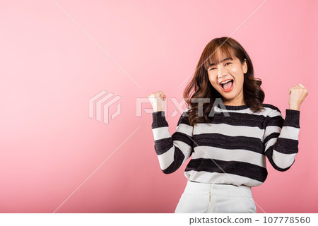 Portrait of a jubilant Asian woman with raised fists, celebrating her success with a happy expression. Studio shot isolated on a pink background, conveying victory and joy. 107778560