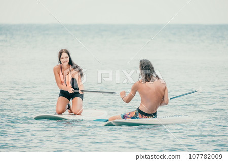 Sea woman and man on sup. Silhouette of happy young woman and man, surfing on SUP board, confident paddling through water surface. Idyllic sunset. Active lifestyle at sea or river. Sea woman and man on sup. Silhouette of happy young woman and man, surfing on SUP board, confident paddling through water surface. Idyllic sunset. Active lifestyle at sea or river. 107782009