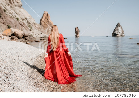 Woman travel sea. Happy tourist in red dress enjoy taking picture outdoors for memories. Woman traveler posing on the rock at sea bay surrounded by volcanic mountains, sharing travel adventure journey Woman travel sea. Happy tourist in red dress enjoy taking picture outdoors for memories. Woman traveler posing on the rock at sea bay surrounded by volcanic mountains, sharing travel adventure journey 107782010