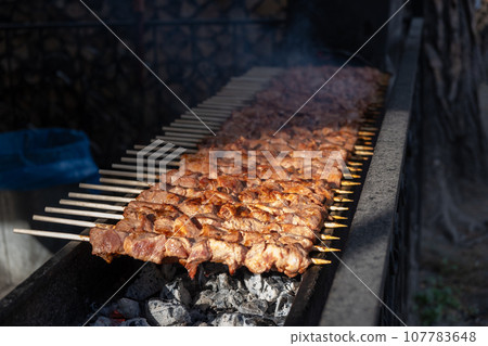 A lot of juicy meat kebabs in a row on the grill. Meat pieces strung on wooden skewers on the grill. The process of cooking kebabs with a lot of smoke. Cooking in nature A lot of juicy meat kebabs in a row on the grill. Meat pieces strung on wooden skewers on the grill. The process of cooking kebabs with a lot of smoke. Cooking in nature 107783648