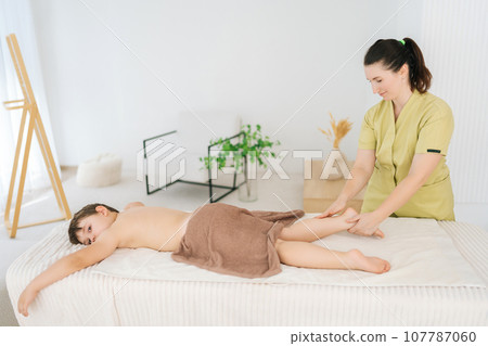 Side view of five year old boy lying on massage table having lymphatic drainage massage of legs and lower legs. Portrait of female massage therapist massaging legs of little kid client in spa salon. Side view of five year old boy lying on massage table having lymphatic drainage massage of legs and lower legs. Portrait of female massage therapist massaging legs of little kid client in spa salon. 107787060