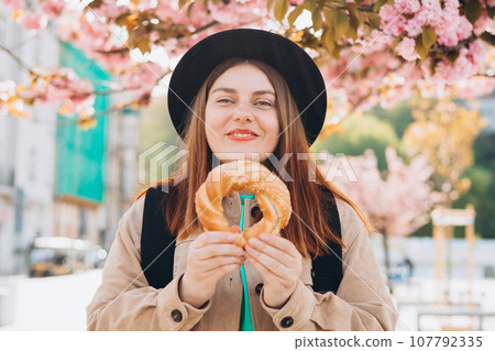 Attractive young female tourist is holding pretzel, traditional polish or turkish bagel. Traveling Europe in spring. High quality photo 107792335