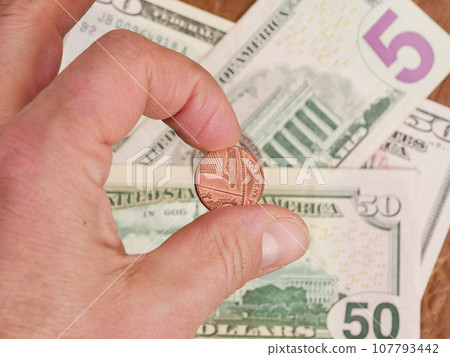 A man's hand holds a coin close-up on the background of paper dollars. A man's hand holds a coin close-up on the background of paper dollars. 107793442