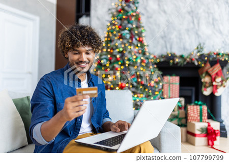 Man is sitting on a sofa in the living room of the house for Christmas, hispanic smiling, satisfied, choosing gifts and goods online in an online store, holding a bank credit card near tree. Man is sitting on a sofa in the living room of the house for Christmas, hispanic smiling, satisfied, choosing gifts and goods online in an online store, holding a bank credit card near tree. 107799799