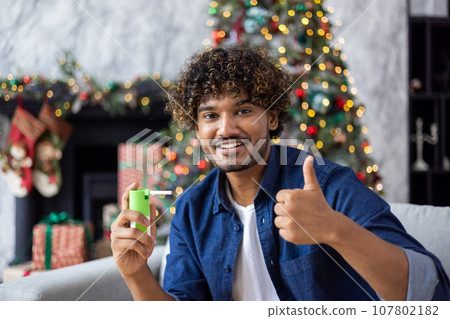 A man uses virtual reality glasses for an online video call and remote communication with friends and relatives, sits on sofa in living room, near Christmas tree, celebrates New Year and Christmas. A man uses virtual reality glasses for an online video call and remote communication with friends and relatives, sits on sofa in living room, near Christmas tree, celebrates New Year and Christmas. 107802182