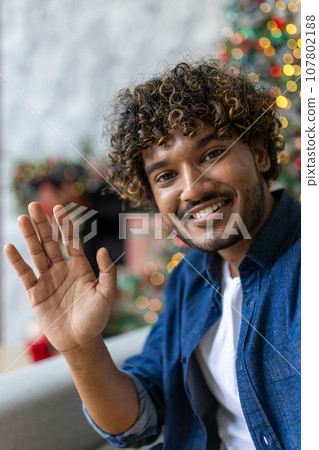 vertical shot man at home looking at phone camera talking remotely with friends and family, happy smiling waving at camera saying hello, sitting on sofa for christmas near tree, celebrating new year vertical shot man at home looking at phone camera talking remotely with friends and family, happy smiling waving at camera saying hello, sitting on sofa for christmas near tree, celebrating new year 107802188
