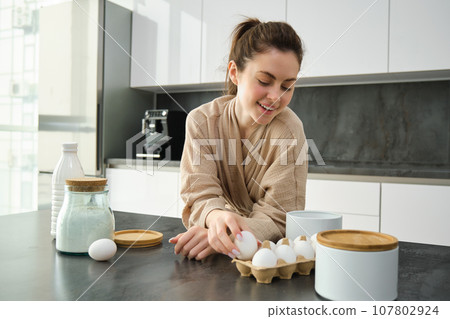 Attractive young cheerful girl baking at the kitchen, making dough, holding recipe book, having ideas Attractive young cheerful girl baking at the kitchen, making dough, holding recipe book, having ideas 107802924