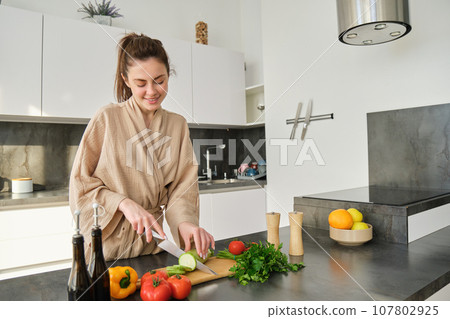 Portrait of good-looking woman cooking salad in the kitchen, chopping vegetables and smiling, preparing healthy meal, leading healthy lifestyle and eating raw food Portrait of good-looking woman cooking salad in the kitchen, chopping vegetables and smiling, preparing healthy meal, leading healthy lifestyle and eating raw food 107802925