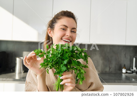 Portrait of beautiful smiling girl with bouquet of parsley, standing in the kitchen and cooking, adding herbs to healthy fresh salad or meal, preparing food Portrait of beautiful smiling girl with bouquet of parsley, standing in the kitchen and cooking, adding herbs to healthy fresh salad or meal, preparing food 107802926