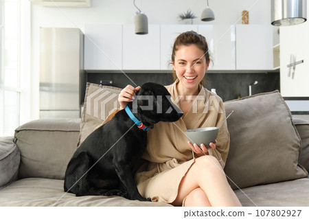 Morning and lifestyle concept. Happy young woman on sofa, sitting with dog, cuddle her puppy and eating cereals, having breakfast, laughing and smiling Morning and lifestyle concept. Happy young woman on sofa, sitting with dog, cuddle her puppy and eating cereals, having breakfast, laughing and smiling 107802927
