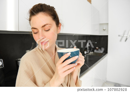 Portrait of happy, smiling modern woman, starts her day with morning mug of tea. Girl drinking coffee in the kitchen, standing with mug and looking relaxed 107802928
