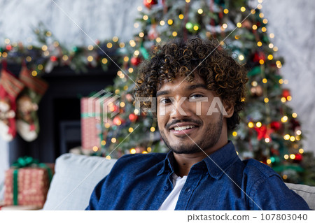 Christmas portrait of young happy man, Hispanic man smiling and looking at camera, happy and celebrating Christmas sitting on sofa in living room near decorated Christmas tree. 107803040