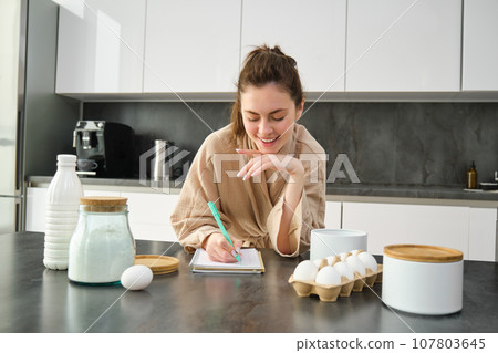 Attractive young cheerful girl baking at the kitchen, making dough, holding recipe book, having ideas Attractive young cheerful girl baking at the kitchen, making dough, holding recipe book, having ideas 107803645