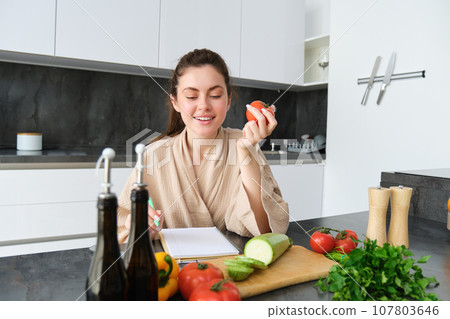 Portrait of beautiful smiling woman, writing her healthy menu, eating tomato while cooking, making grocery list, sitting in the kitchen 107803646