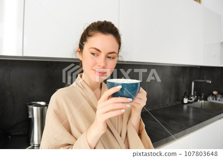 Portrait of happy young woman starts her morning with mug of coffee, drinking tea from cup, standing in the kitchen, smiling cheerfully 107803648