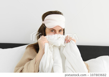 Cute girlfriend with sleeping mask on forehead, cover her face behind duvet and giggle, looking playful and flirty at camera, resting in bedroom on her cosy bed with white sheets Cute girlfriend with sleeping mask on forehead, cover her face behind duvet and giggle, looking playful and flirty at camera, resting in bedroom on her cosy bed with white sheets 107803682