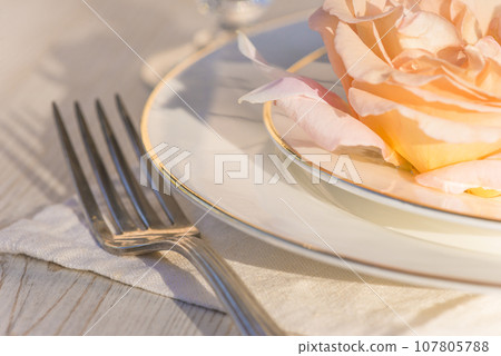 Romantic table setting; close up for salmon rose flower on the white plates, fork on the white napkin son sunlight Romantic table setting; close up for salmon rose flower on the white plates, fork on the white napkin son sunlight 107805788