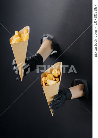 Hands holding French fries through torn holes in black paper background. Street food concept 107805867