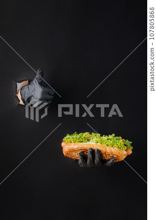 Hand holding hot dog and showing a thumb up through torn holes in black paper background. Street food concept Hand holding hot dog and showing a thumb up through torn holes in black paper background. Street food concept 107805868