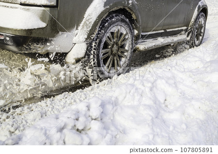 Road snow flies up from a vehicle's spinning wheel. Car's wheels spin and spew up pieces of snow it attempts to gain traction on the slippery road. 107805891