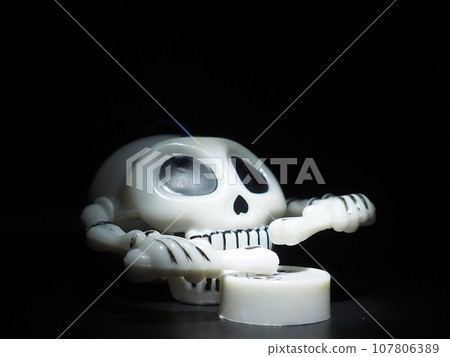 Skull with drumsticks and drum. Terrible clockwork toy. The skeleton is made of the skull, bones, hands and fingers. Black background. Halloween and horror theme. White skull with black eye sockets. 107806389