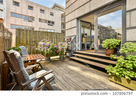 a wooden deck with chairs and potted plants in the foreground, on a sunny day at an apartment building 107806797