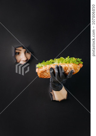 Female hand holding hot dog and looks through holes in black paper background. Street food concept 107806850