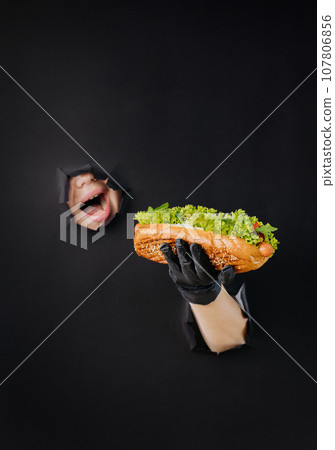 Female hand holding hot dog and looks through holes in black paper background. Street food concept 107806856