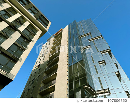 Fragment of modern tall building with glass facade against blue sky. High growing buildings low angle view. Modern multi-story standstill in residential neighborhood. Condominium, apartments for sale. 107806860