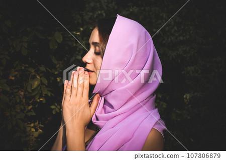Beautiful young woman with head covered with silk purple scarf is standing against green natural background. Devout girl praying with palms folded together. Spirituality, faith, religion, gratitude. 107806879