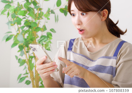 A woman in an apron holding a smartphone with a surprised expression 107808339