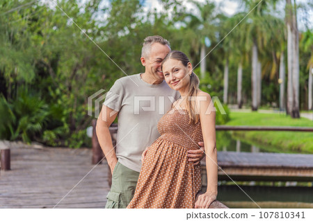A happy, mature couple over 40, enjoying a leisurely walk in a park, their joy evident as they embrace the journey of pregnancy later in life A happy, mature couple over 40, enjoying a leisurely walk in a park, their joy evident as they embrace the journey of pregnancy later in life 107810341