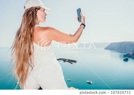 A woman cyclist on a mountain bike looking at the landscape of mountains and sea. Rear view of cyclist woman standing in front to the sea with outstretched arms. Freedom and healthy lifestyle concept. 107810731