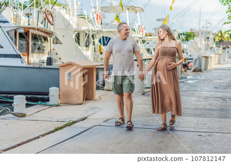 A happy, mature couple over 40, enjoying a leisurely walk on the waterfront, their joy evident as they embrace the journey of pregnancy later in life A happy, mature couple over 40, enjoying a leisurely walk on the waterfront, their joy evident as they embrace the journey of pregnancy later in life 107812147