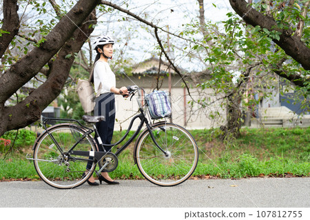 A young woman wearing a cycling helmet and pulling her bicycle along the riverside promenade A young woman wearing a cycling helmet and pulling her bicycle along the riverside promenade 107812755