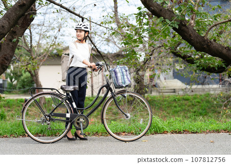 A young woman wearing a cycling helmet and pulling her bicycle along the riverside promenade A young woman wearing a cycling helmet and pulling her bicycle along the riverside promenade 107812756