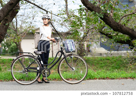 一位年輕女子戴著自行車頭盔,沿著河濱長廊拉著自行車 一位年輕女子戴著自行車頭盔,沿著河濱長廊拉著自行車 107812758