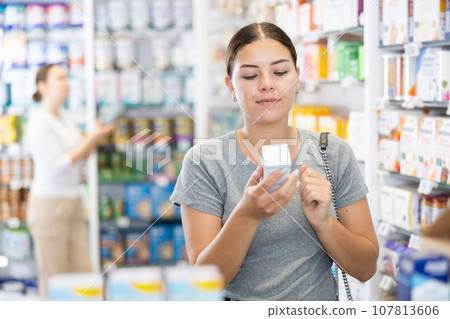 In pharmacy, woman carefully examines package of cream for problem skin 107813606
