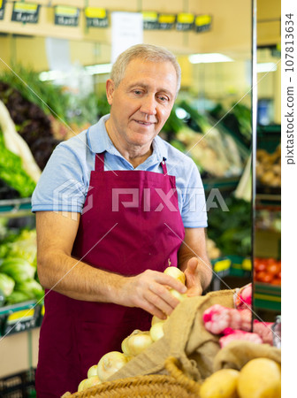 Aged salesman putting onions on food stall in greengrocery 107813634