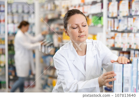 Adult female pharmacist arranges products in pharmacy 107813640