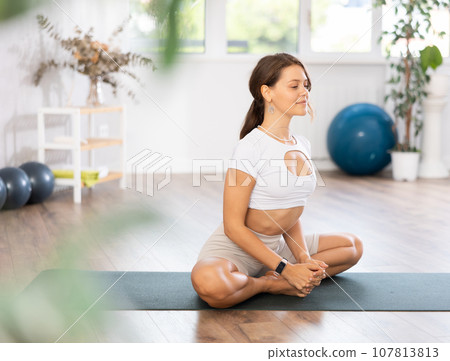 Young woman yoga trainer performing lotus pose variation on black rug 107813813
