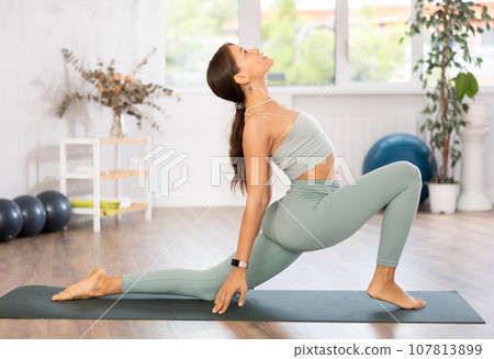 Young woman yoga trainer performing crescent lunge pose on black rug Young woman yoga trainer performing crescent lunge pose on black rug 107813899
