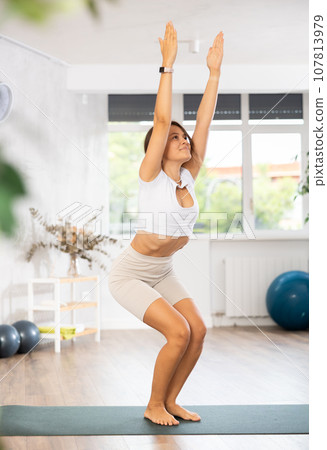 Young woman yoga trainer performing chair pose on black rug Young woman yoga trainer performing chair pose on black rug 107813979