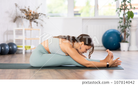 Young woman yoga trainer performing seated forward bend pose on black rug Young woman yoga trainer performing seated forward bend pose on black rug 107814042